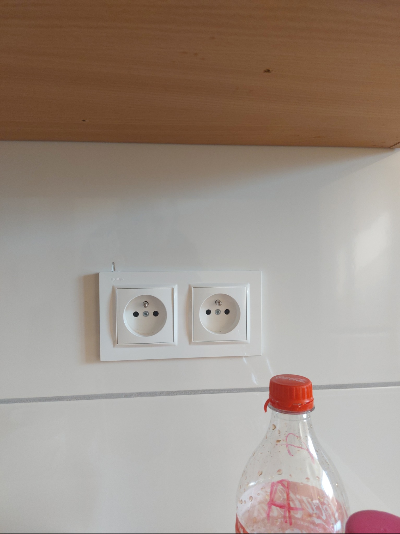 Białe, podwójne gniazdko elektryczne marki Simon zamontowane na białej ścianie w kuchni, widoczne spod szafki, z butelką plastikową w dolnym rogu.