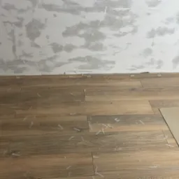 Układanie podłogi z płytek imitujących drewno, widoczne krzyżyki dystansowe i czerwone kliny przy ścianie, ściana przygotowana do dalszych prac wykończeniowych.