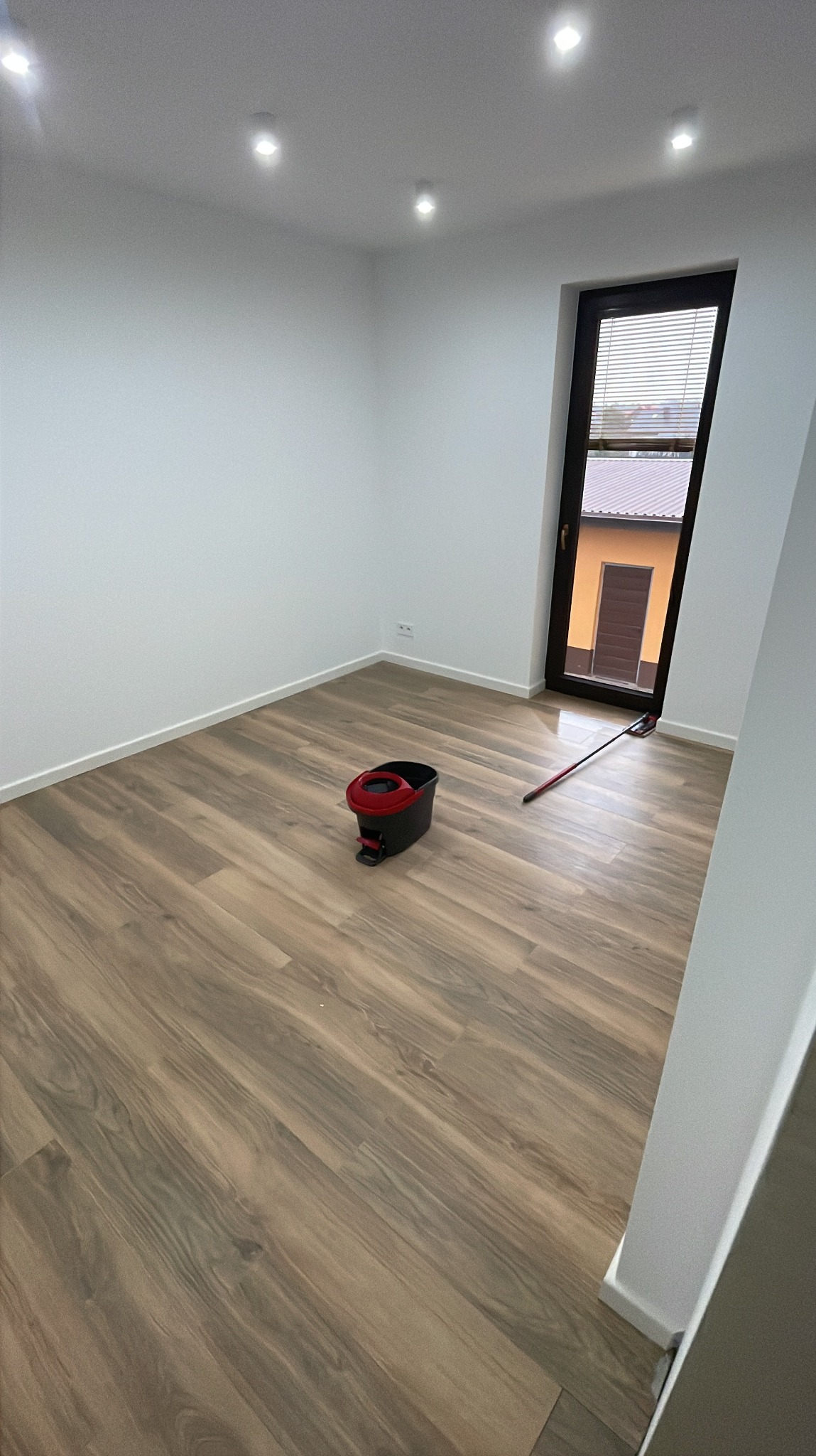 Pokój po remoncie z panelami podłogowymi i mopem. Widok na okno z żaluzjami. Świeżo wykończone wnętrze z oświetleniem punktowym.