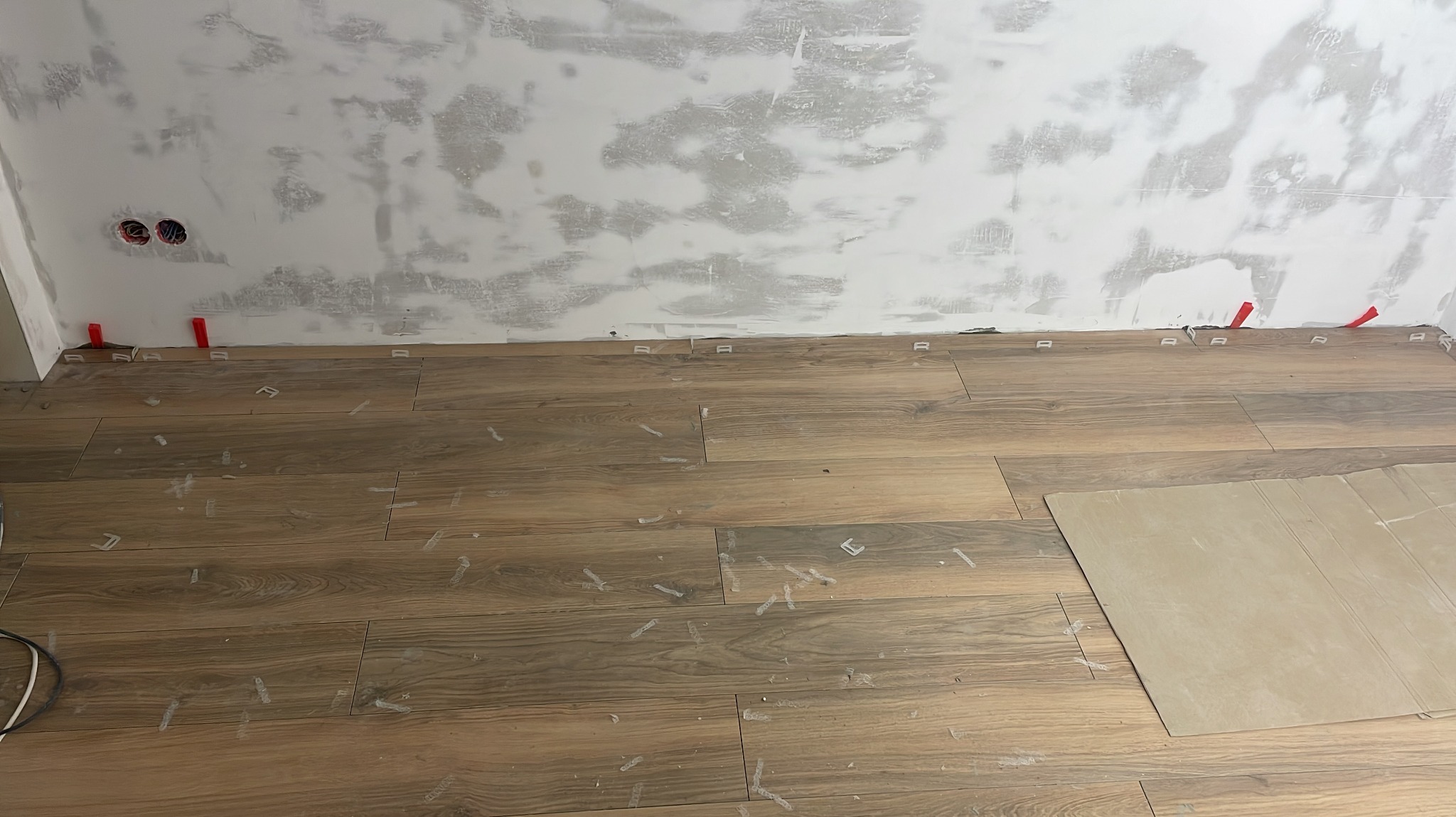 Układanie podłogi z płytek imitujących drewno, widoczne krzyżyki dystansowe i czerwone kliny przy ścianie, ściana przygotowana do dalszych prac wykończeniowych.