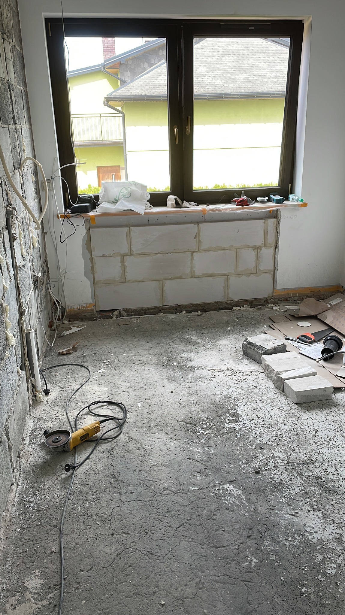 Wnętrze w trakcie prac murarskich: ściana z białych bloczków pod oknem, narzędzia na parapecie, szlifierka kątowa na szarym podłożu. Widok na budynek z zewnątrz.