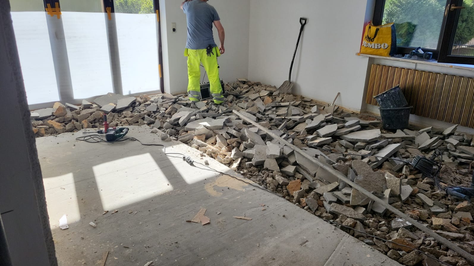 Demontaż podłogi w trakcie remontu mieszkania: gruz, narzędzia (wiertarka, łopata), pracownik w odblaskowych spodniach. Widoczne okno i kaloryfer.