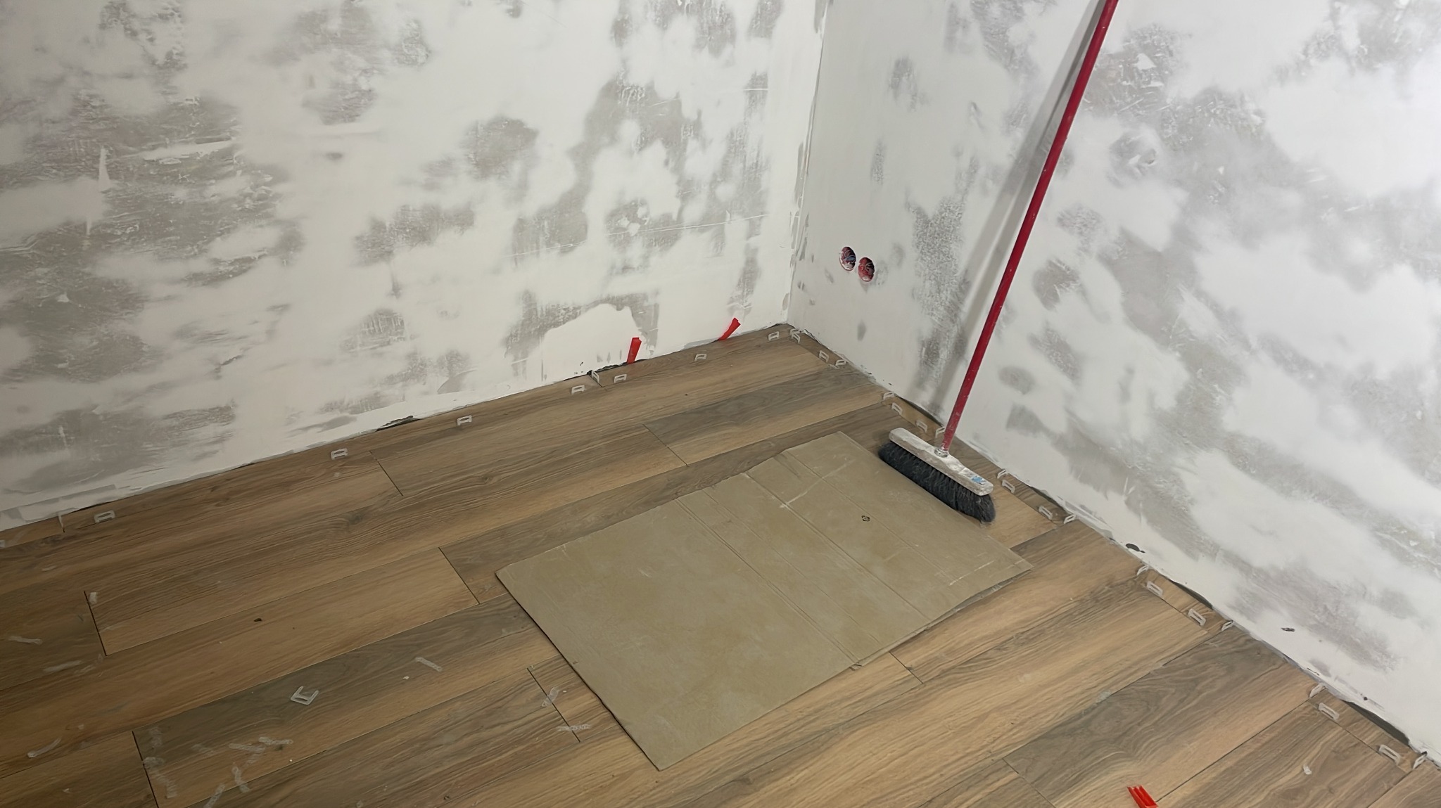 Układanie drewnopodobnych płytek podłogowych w rogu pomieszczenia. Widoczne klipsy poziomujące, szpachlowane ściany, karton ochronny i szczotka.