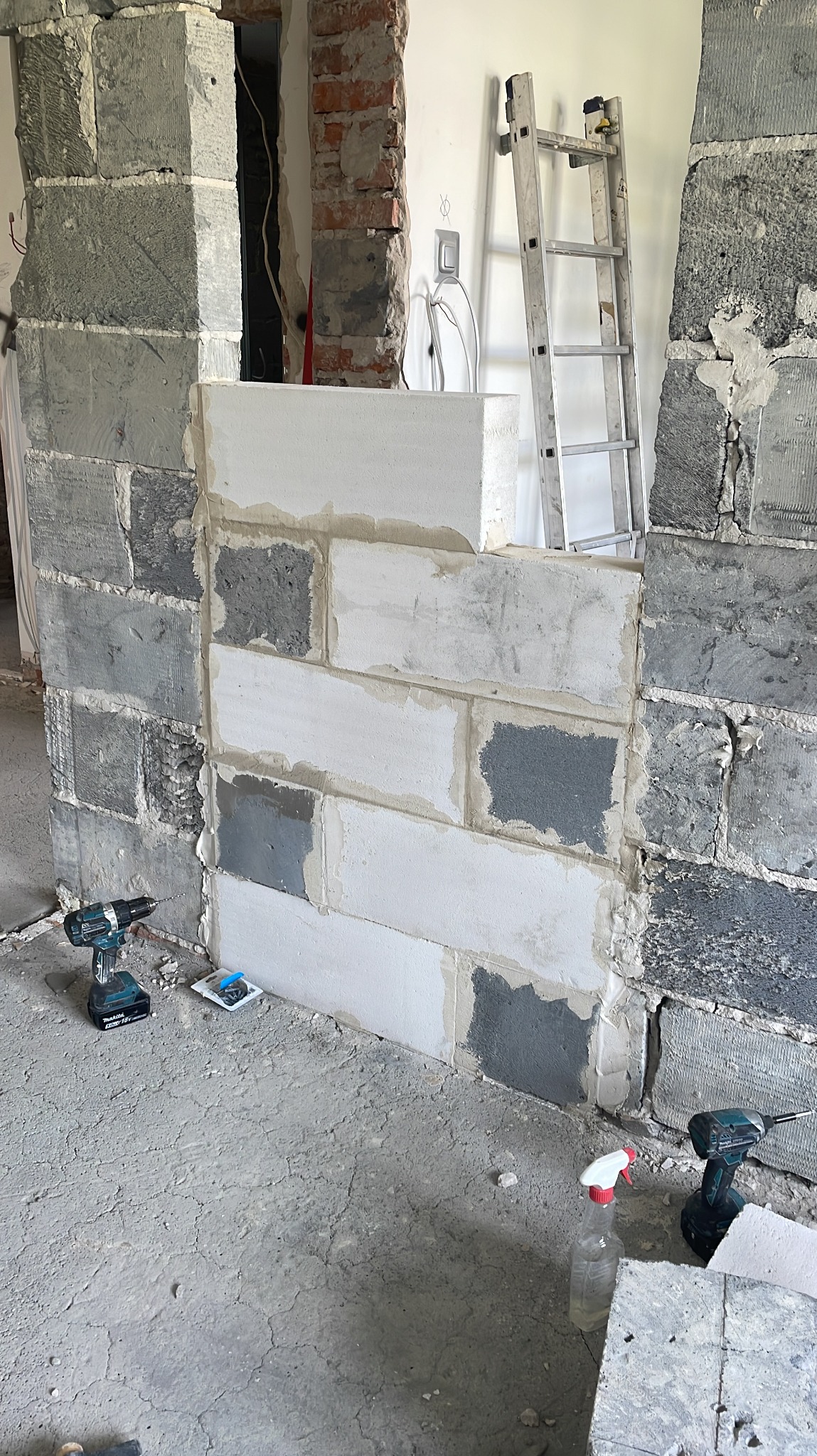 Murowanie ściany z bloczków z betonu komórkowego, widoczne narzędzia (wiertarki) i drabina oparta o ścianę. Prace murarskie w trakcie remontu mieszkania.
