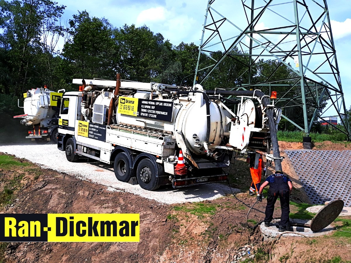 Specjalistyczny pojazd do czyszczenia kanalizacji w trakcie pracy przy studzience, w tle linia wysokiego napięcia i drugi pojazd. Widoczni pracownicy w kombinezonach.