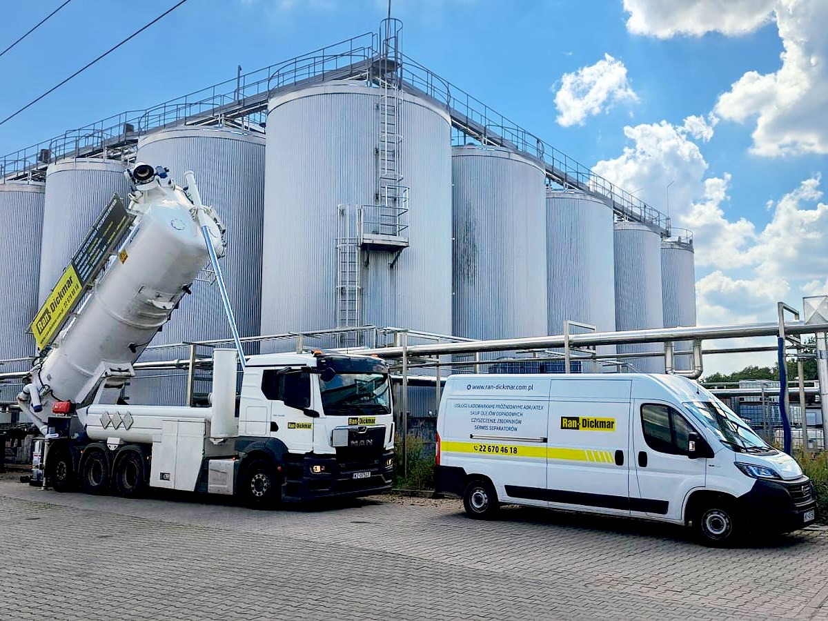 Specjalistyczny pojazd do czyszczenia zbiorników przemysłowych na tle silosów i błękitnego nieba. Widoczny również biały bus serwisowy z logo firmy.