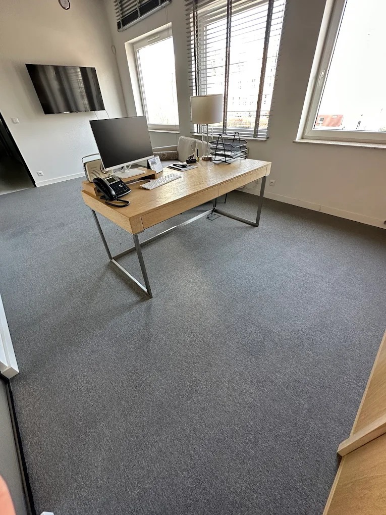 Jasne biuro po sprzątaniu: drewniane biurko z komputerem, telefonem, lampką i dokumentami na szarym dywanie pod oknami z żaluzjami. Telewizor na ścianie.