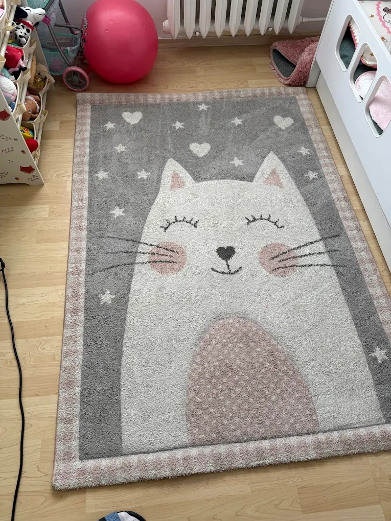 Dziecięcy dywan z motywem kota na drewnianej podłodze, po praniu. Widoczne serduszka i gwiazdki we wzorze. Jasne, pastelowe kolory.