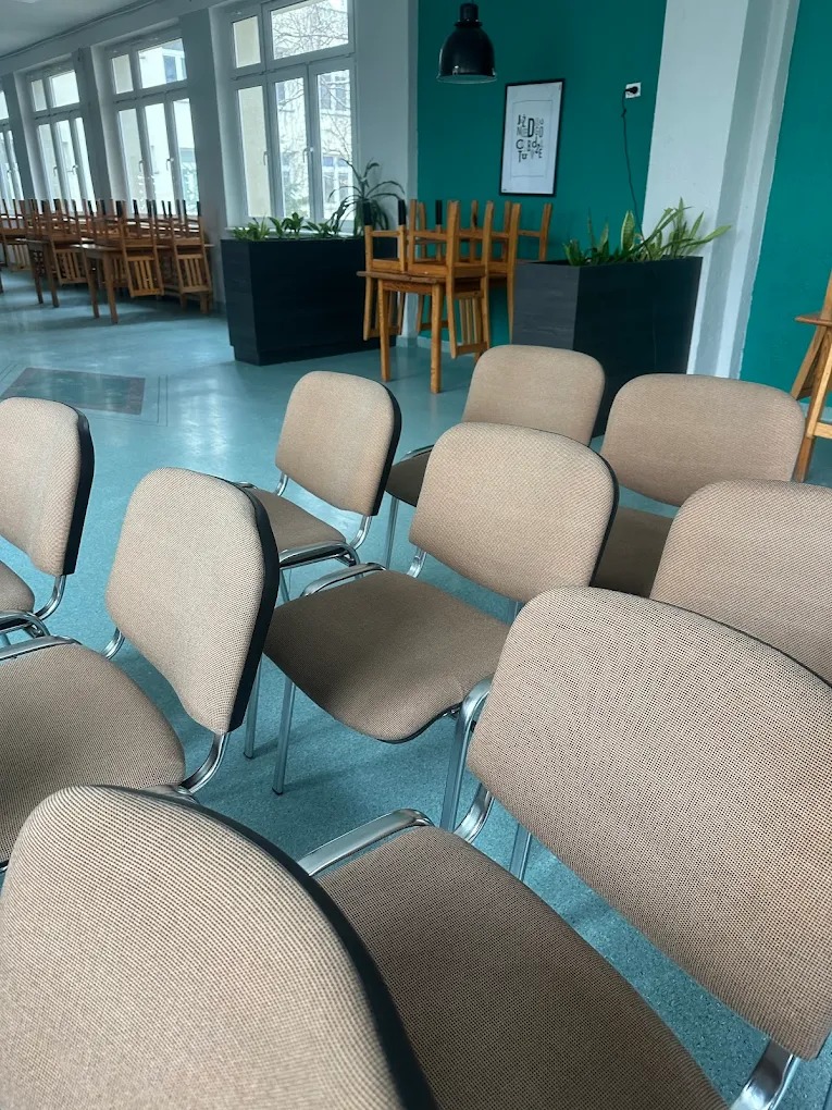 Rząd krzeseł tapicerowanych w sali konferencyjnej, z zieloną ścianą w tle i naturalnym światłem wpadającym przez okna. Widoczne detale tapicerki i metalowe ramy krzeseł.