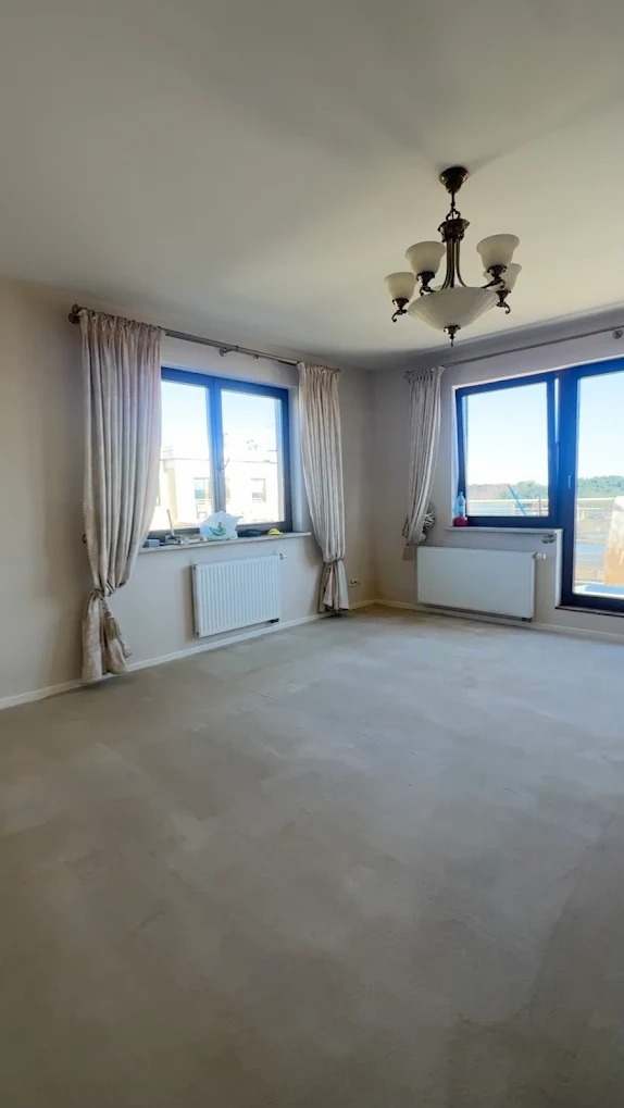 Puste wnętrze pokoju z dwoma oknami z niebieskimi ramami, grzejnikami i kremowymi zasłonami. Na suficie wisi ozdobny, biały żyrandol.