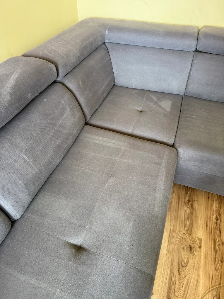 Szary narożnik po praniu: odświeżona tapicerka, brak plam i zapachów. Widoczne ślady wilgoci po czyszczeniu. Parkiet w tle. Ujęcie z bliska, prezentujące efekt usługi.