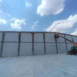 Budowa ściany z betonu komórkowego z użyciem dźwigu teleskopowego JCB, widok na konstrukcję, niebo z chmurami w tle. Prace budowlane w toku.