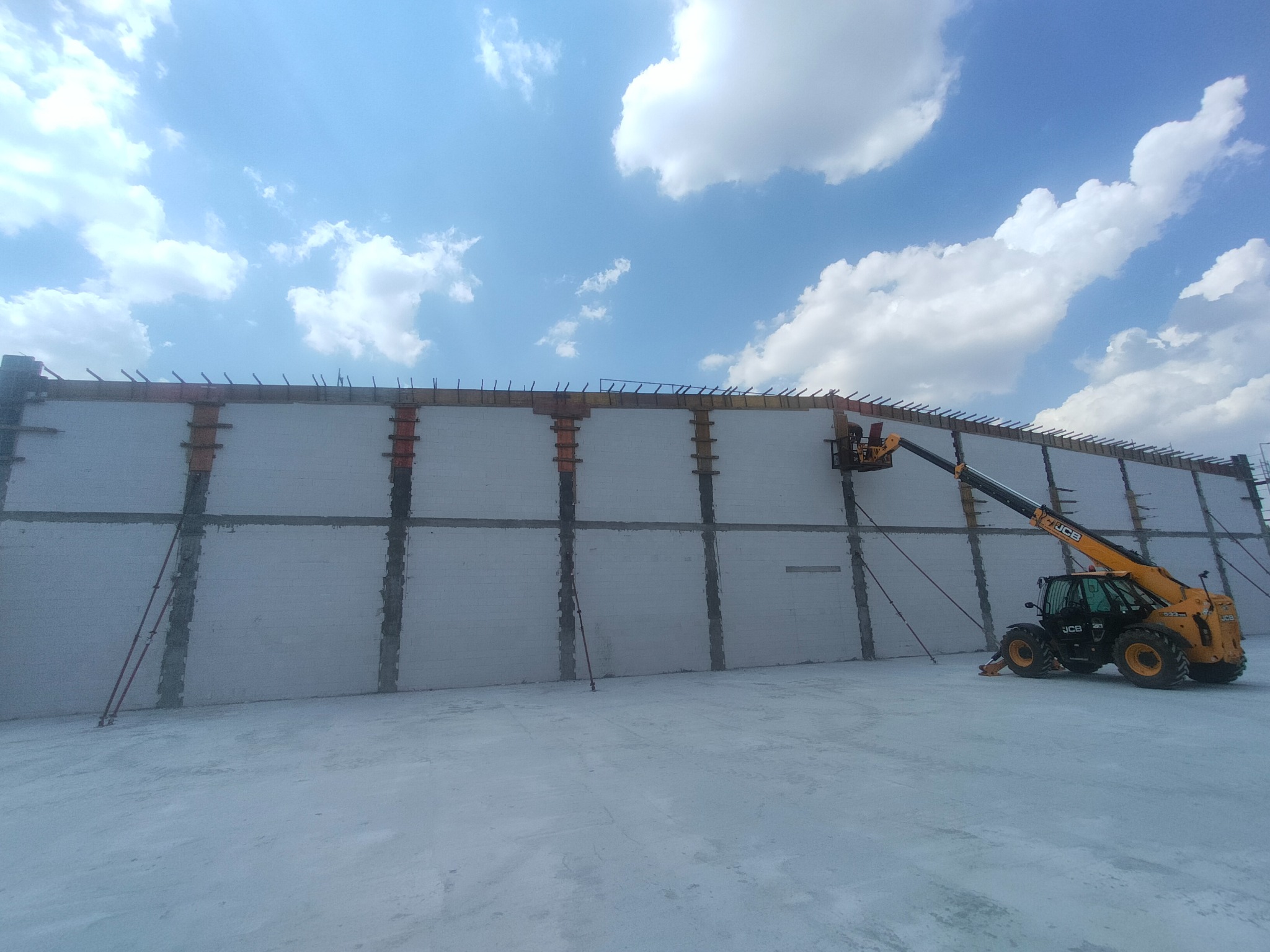 Budowa ściany z betonu komórkowego z użyciem dźwigu teleskopowego JCB, widok na konstrukcję, niebo z chmurami w tle. Prace budowlane w toku.