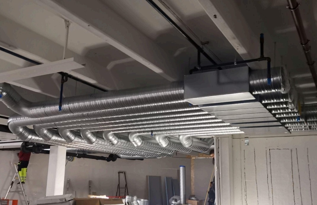 Srebrne, elastyczne przewody wentylacyjne zamontowane pod sufitem. Widoczny fragment instalacji i pracownik na drabinie.