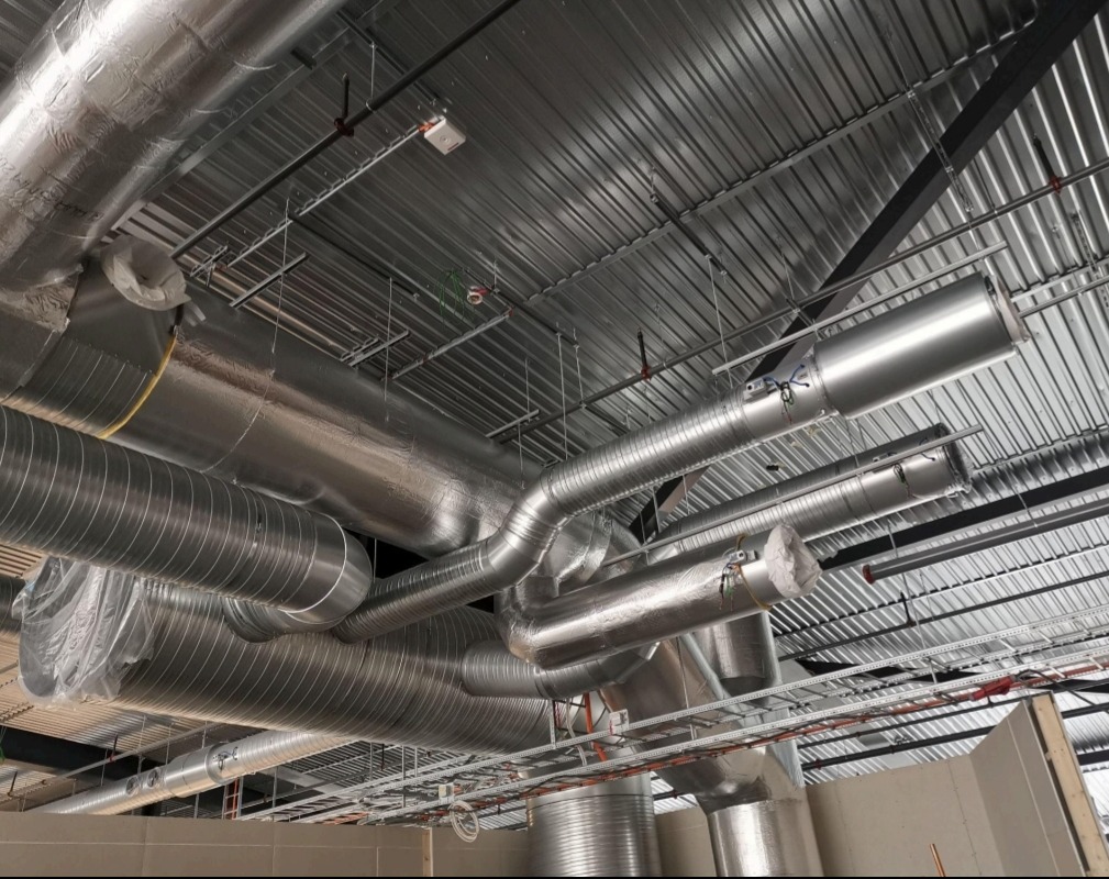Kompleksowa instalacja wentylacyjna z metalowymi rurami pod sufitem, widoczne przewody izolowane i nieizolowane, elementy montażowe i konstrukcja stropu.