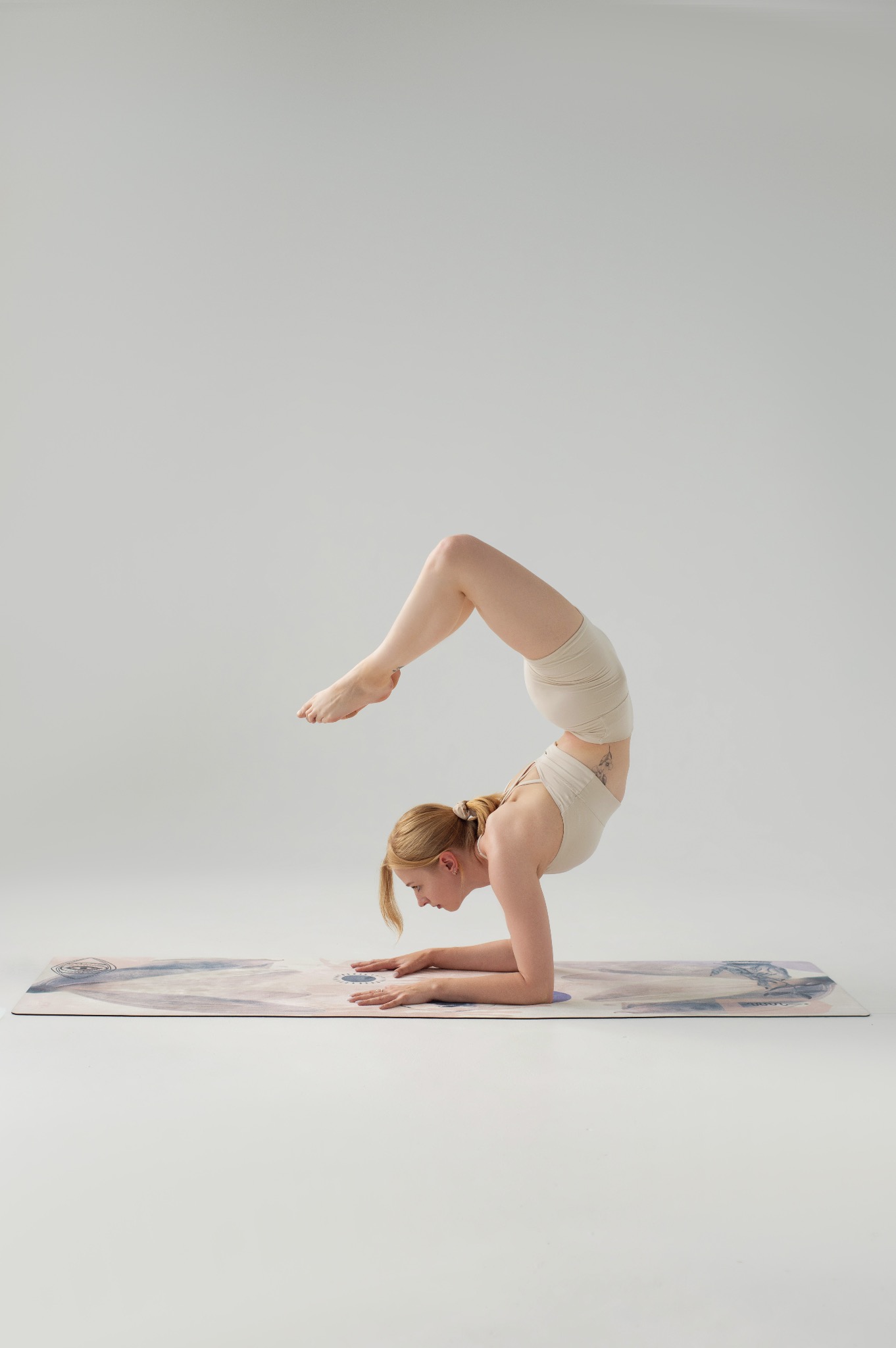 Kobieta w beżowym stroju wykonuje zaawansowaną pozycję jogi, stając na przedramionach na wzorzystej macie. Jasne tło, stonowana kolorystyka, widoczny tatuaż na boku.