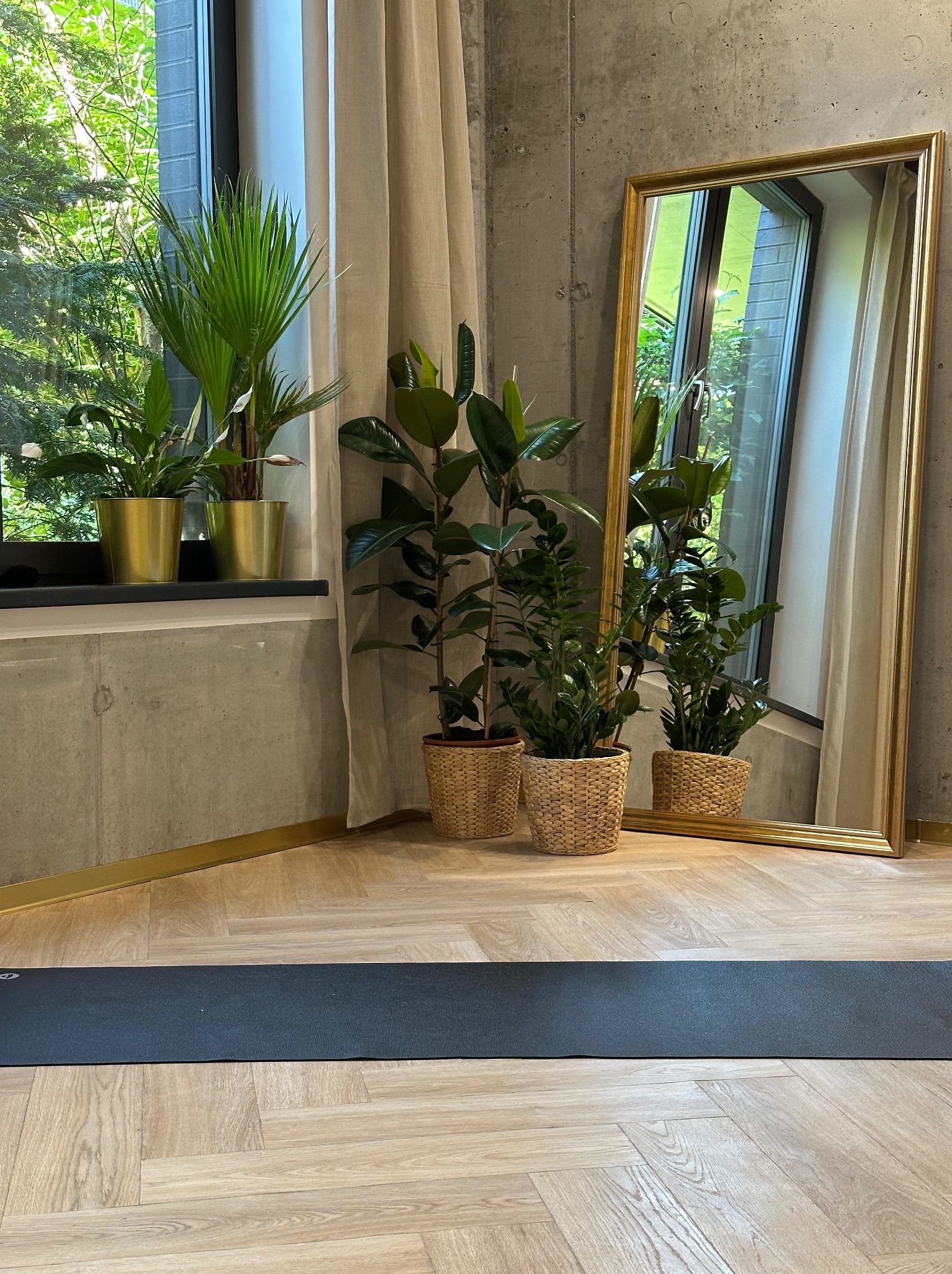 Sala do ćwiczeń z matą do jogi, lustrem w złotej ramie i roślinami doniczkowymi w wiklinowych osłonkach, przy oknie z widokiem na zieleń.