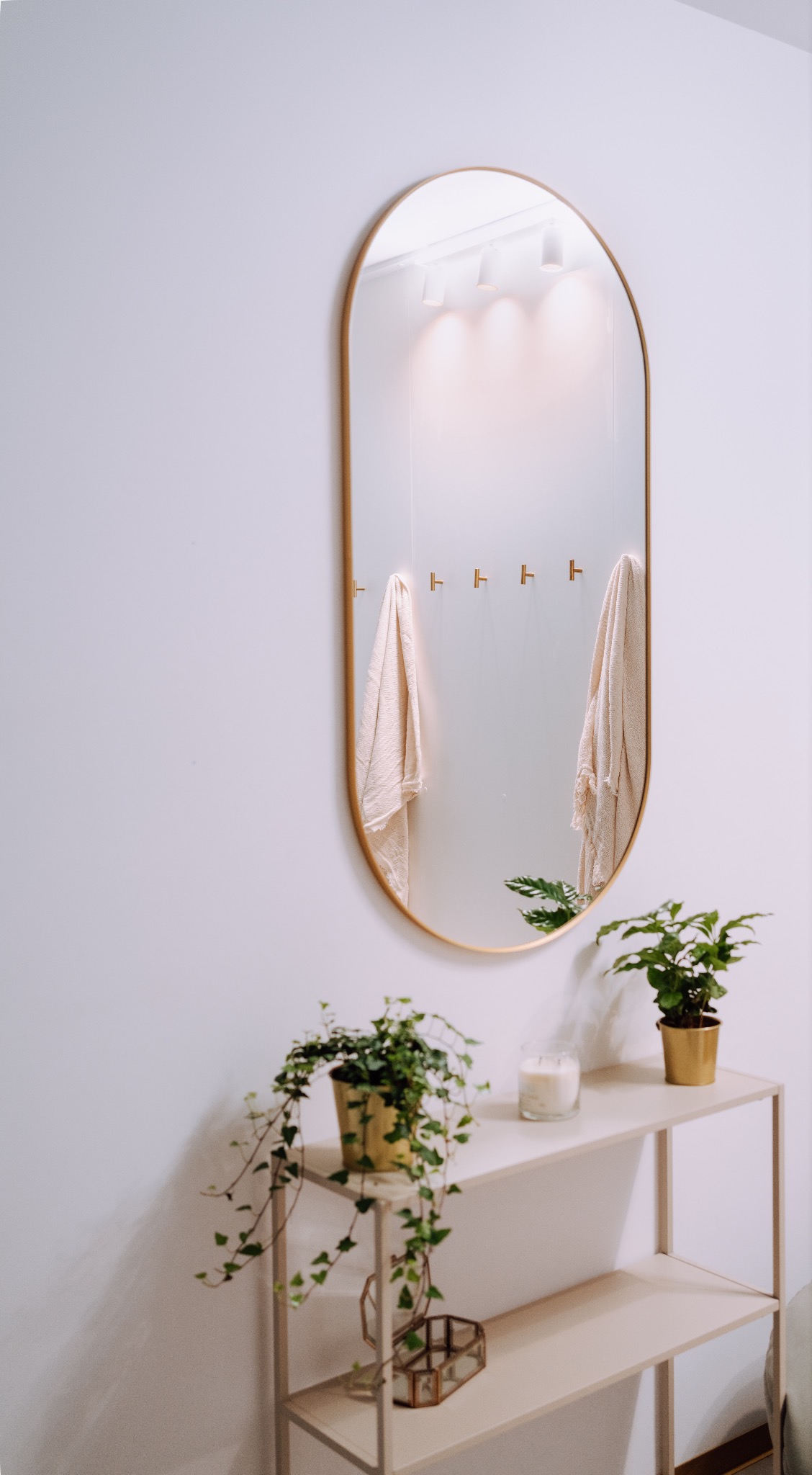 Jasne wnętrze: owalne lustro z złotą ramą, w odbiciu wieszaki i ręczniki. Na półce rośliny w złotych doniczkach i świeca. Minimalistyczny wystrój.