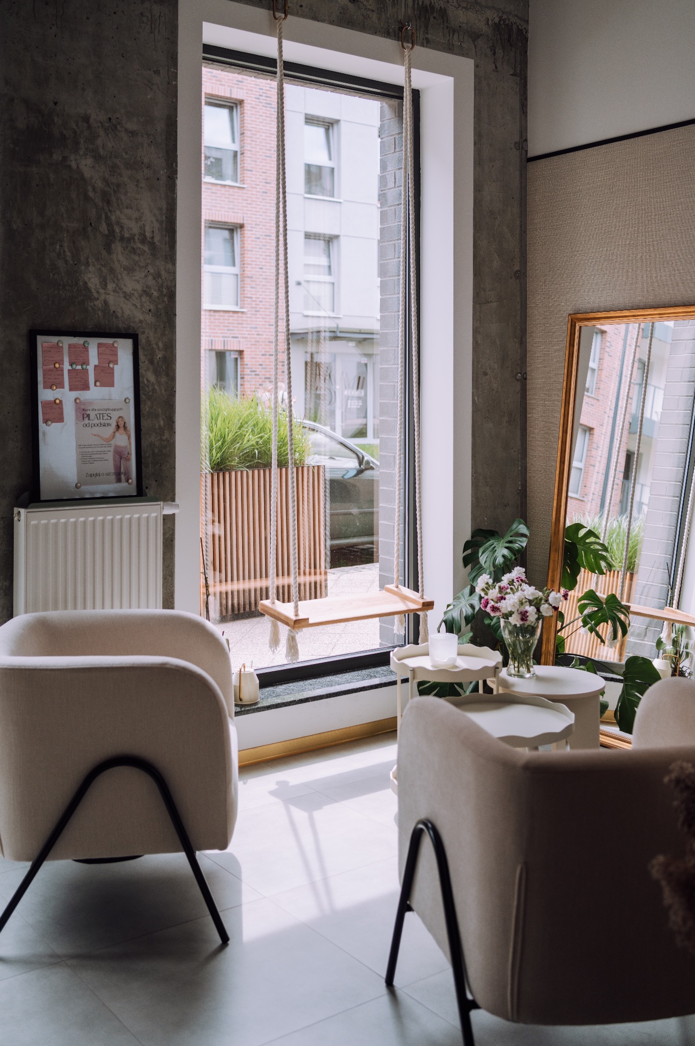 Jasne wnętrze studia Pilates z huśtawką przy oknie, plakat z ofertą zajęć i lustro w złotej ramie. Minimalistyczny wystrój z roślinami i stolikami.