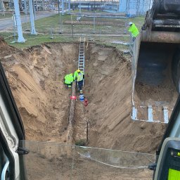 DK-tech - Głęboki wykop z rurą w piasku, widziany z kabiny koparki. W dole pracują robotnicy w żółtych kamizelkach, a w tle widoczna infrastruktura przemysłowa i drabina.