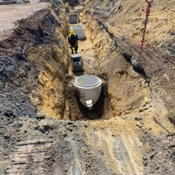 DK-tech - Głęboki wykop z betonowymi kręgami. Robotnik ubija ziemię zagęszczarką. W tle koparka i pracownicy. Widoczne ślady gąsienic na ziemi.