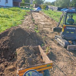DK-tech - Żółta koparka Volvo przygotowuje teren pod drogę na działce. Widoczny wykop, hałdy ziemi i traktor z przyczepą. Prace ziemne w toku.
