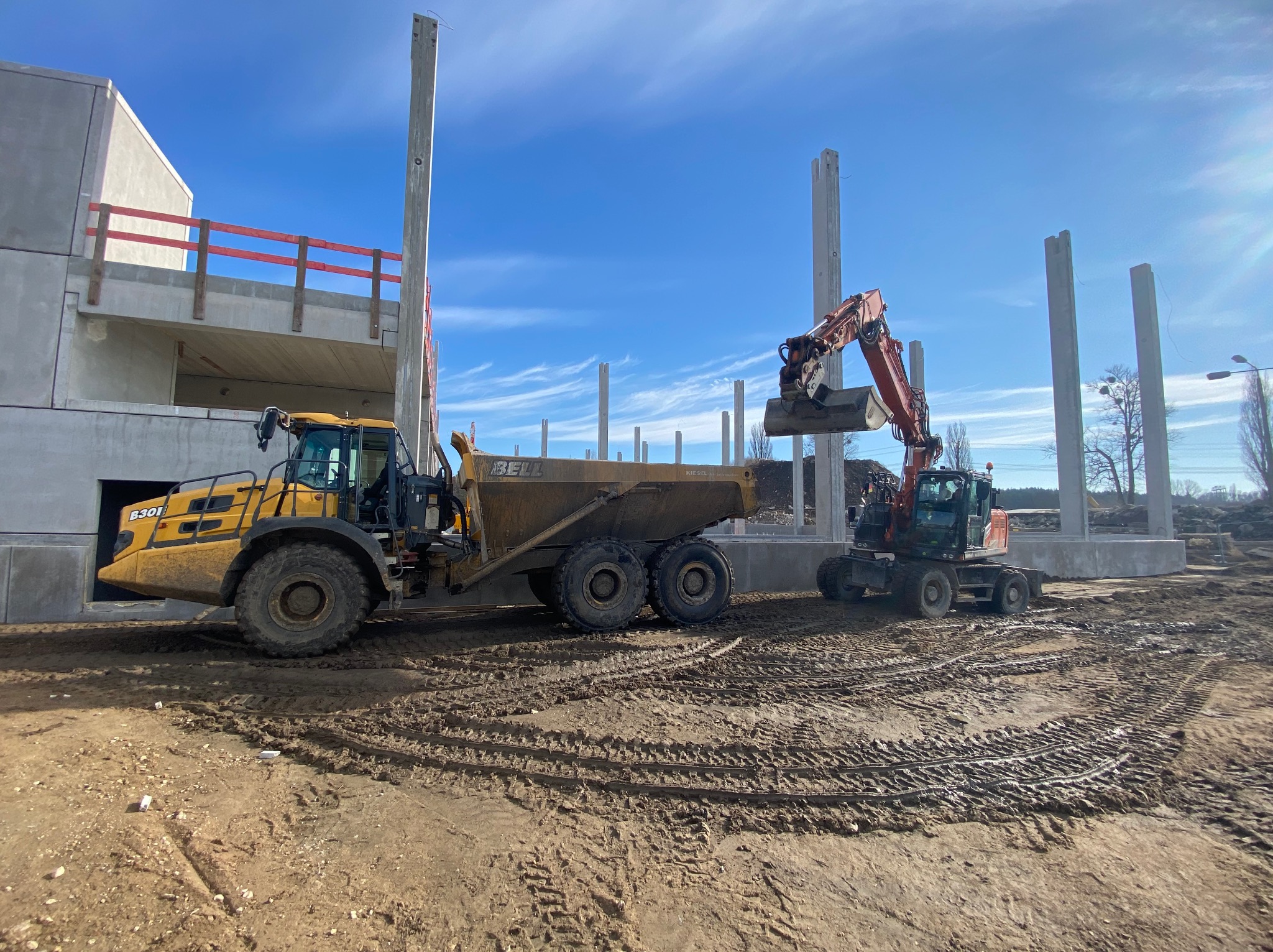 Żółta wywrotka Bell i koparka na placu budowy z betonowymi kolumnami i konstrukcją w tle, pod błękitnym niebem.