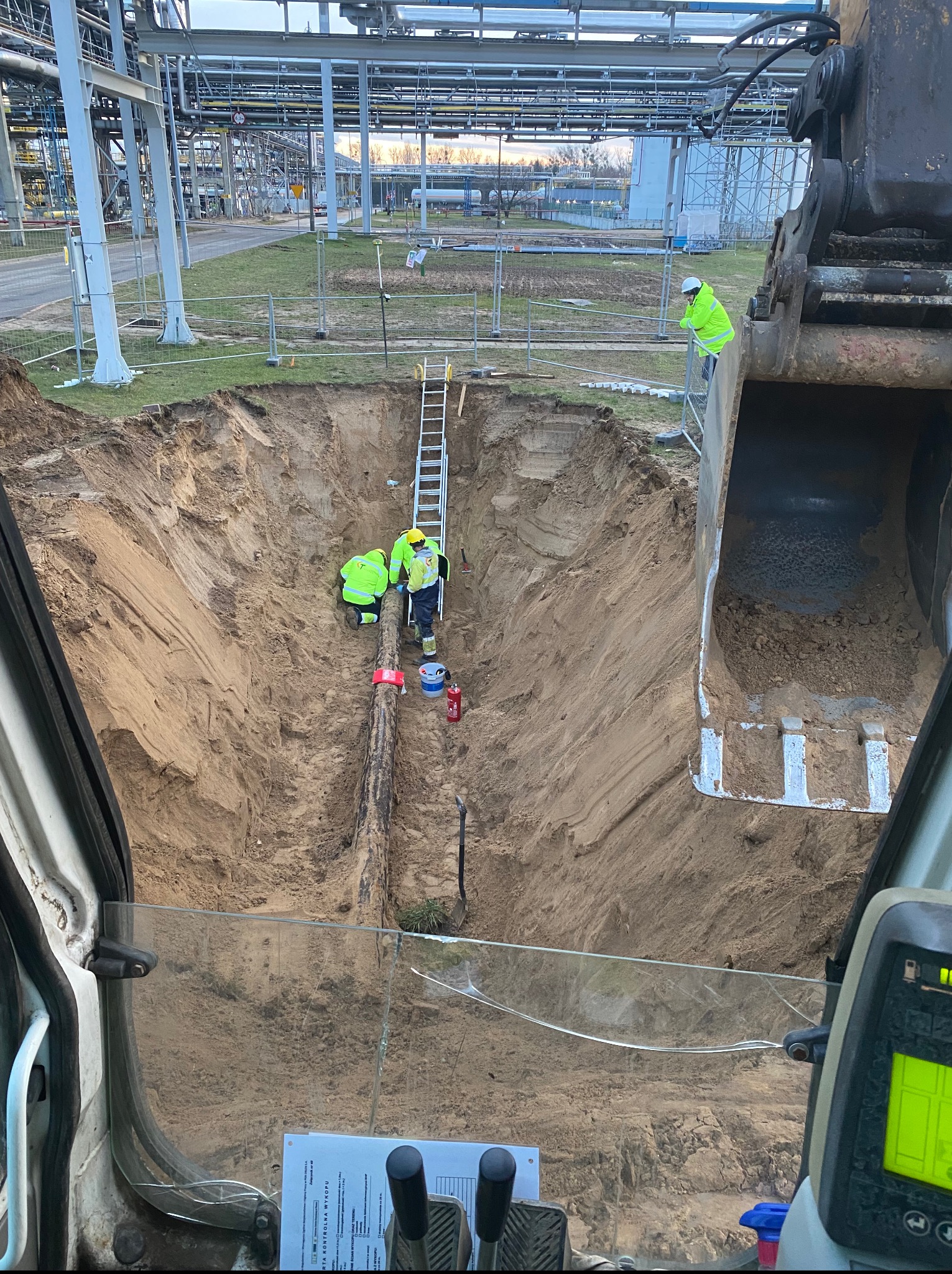 Głęboki wykop z rurą w piasku, widziany z kabiny koparki. W dole pracują robotnicy w żółtych kamizelkach, a w tle widoczna infrastruktura przemysłowa i drabina.