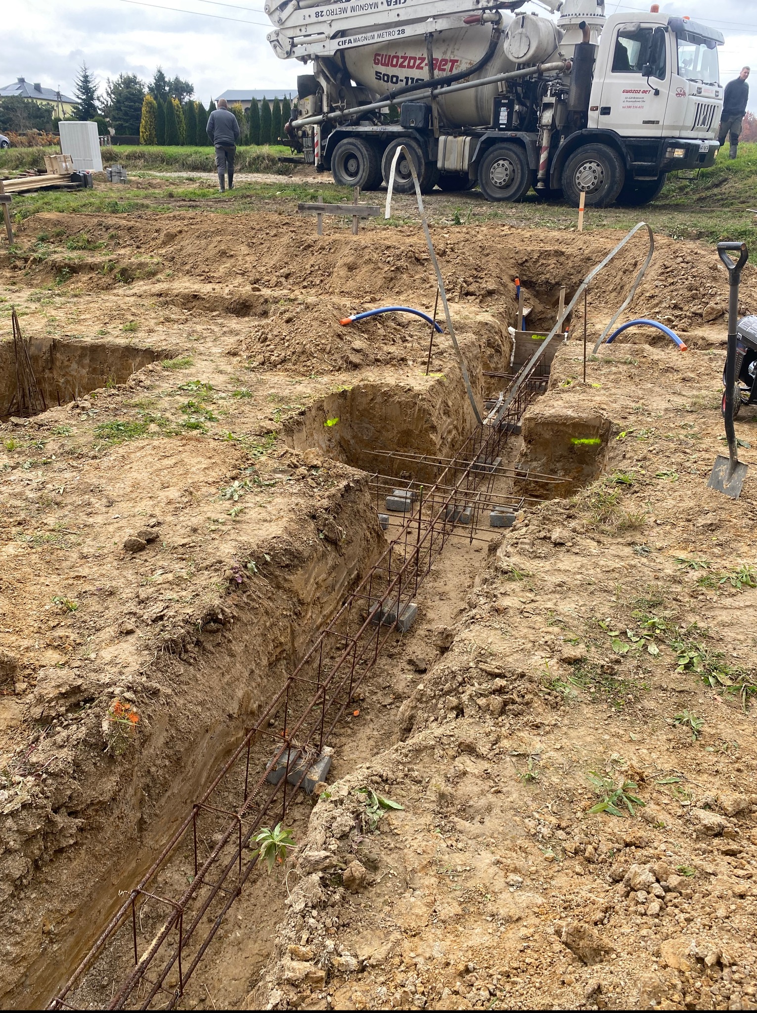 Przygotowanie do wylewki fundamentów: wykopane rowy z ułożonym zbrojeniem, widoczny betonowóz i pracownicy w tle. Etap budowy domu jednorodzinnego.