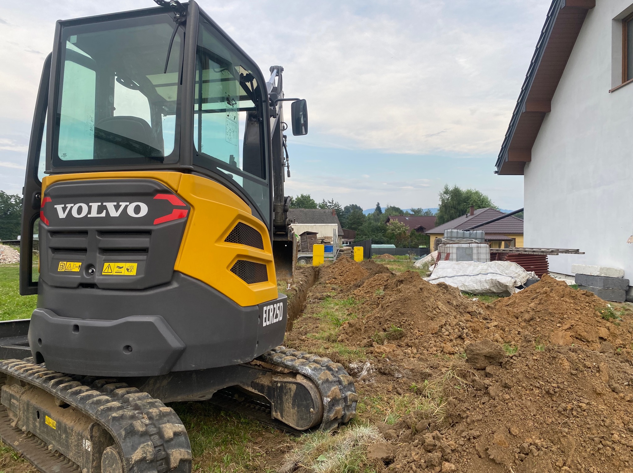 Żółto-czarna koparka Volvo ECR25D w trakcie prac ziemnych na działce, widoczny wykop, hałda ziemi i materiały budowlane. Ujęcie z bliska.