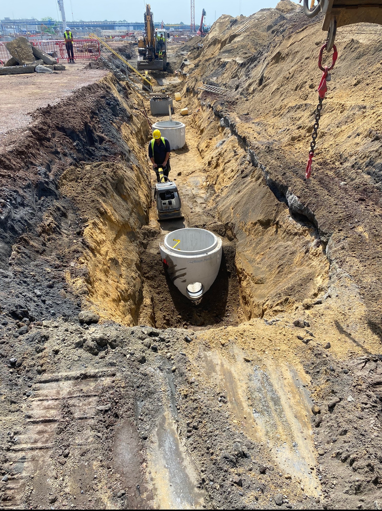 Głęboki wykop z betonowymi kręgami. Robotnik ubija ziemię zagęszczarką. W tle koparka i pracownicy. Widoczne ślady gąsienic na ziemi.