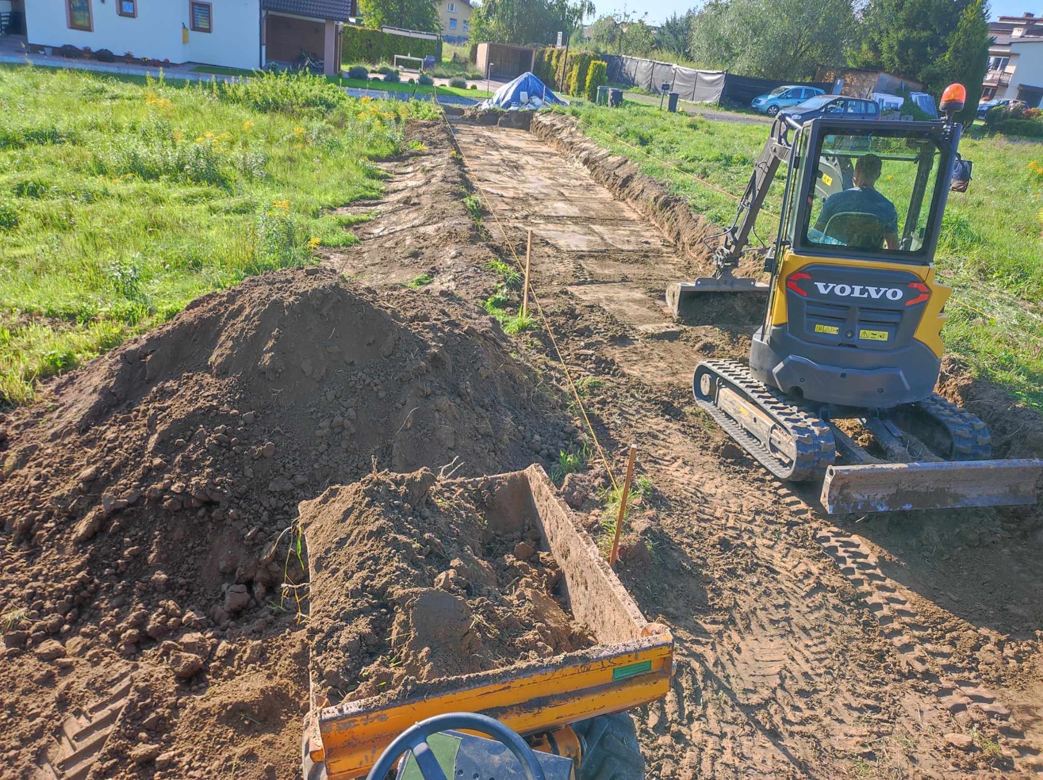Żółta koparka Volvo przygotowuje teren pod drogę na działce. Widoczny wykop, hałdy ziemi i traktor z przyczepą. Prace ziemne w toku.