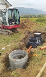 Montaż przydomowej oczyszczalni: betonowy zbiornik z włazem, plastikowy zbiornik, rury, wykop, minikoparka na tle zieleni i domu.