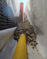 Drenaż liniowy z rurami drenarskimi w wykopie wyłożonym geowłókniną, zasypany żwirem. Widok z dołu wykopu, rury w kolorze pomarańczowym i żółtym.