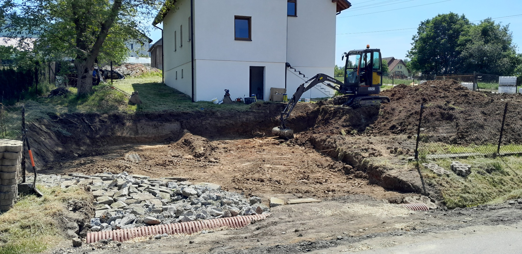 Prace ziemne przy domu jednorodzinnym: wykop z drenażem, gruz, mini koparka Volvo ECR25D w trakcie niwelacji terenu. W tle biały budynek i zieleń.