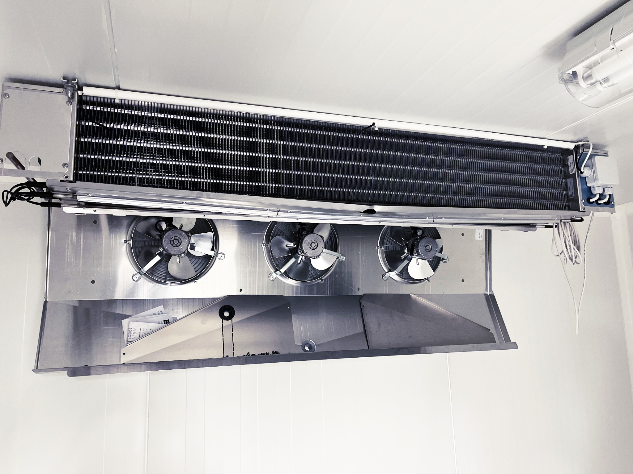 Sufitowa chłodnica przemysłowa z trzema wentylatorami, zamontowana w chłodni. Widoczne przewody i oświetlenie. Ujęcie z dołu, podkreślające detale konstrukcji.