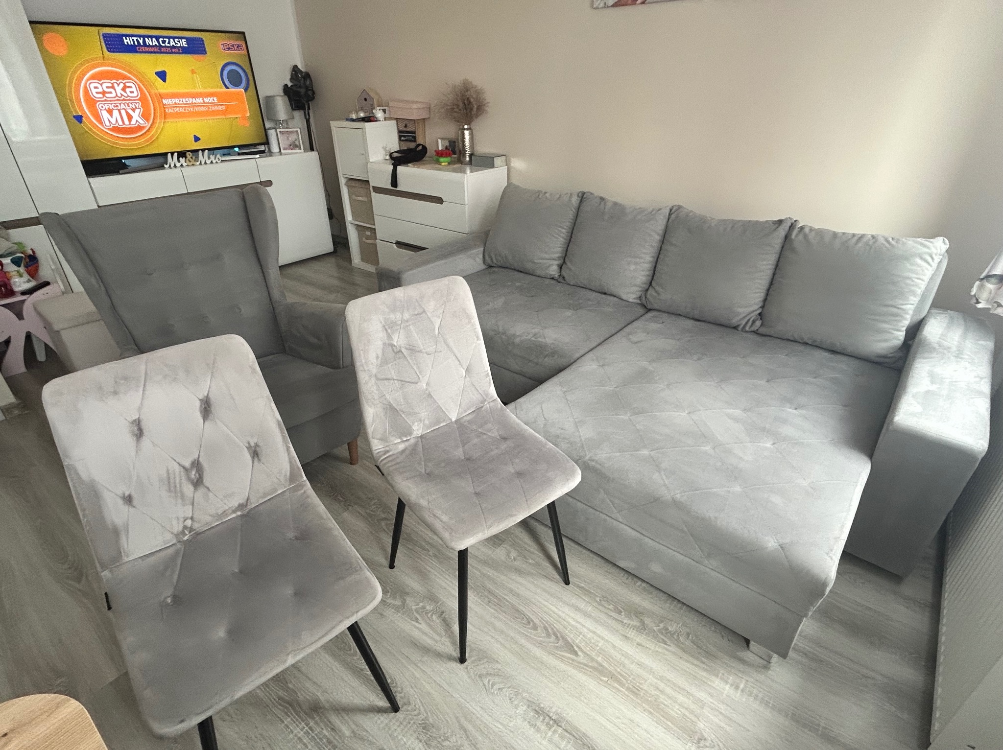 Szary narożnik i krzesła z tapicerką welurową w salonie. Widoczne ślady po czyszczeniu na siedzisku krzesła. Telewizor w tle.