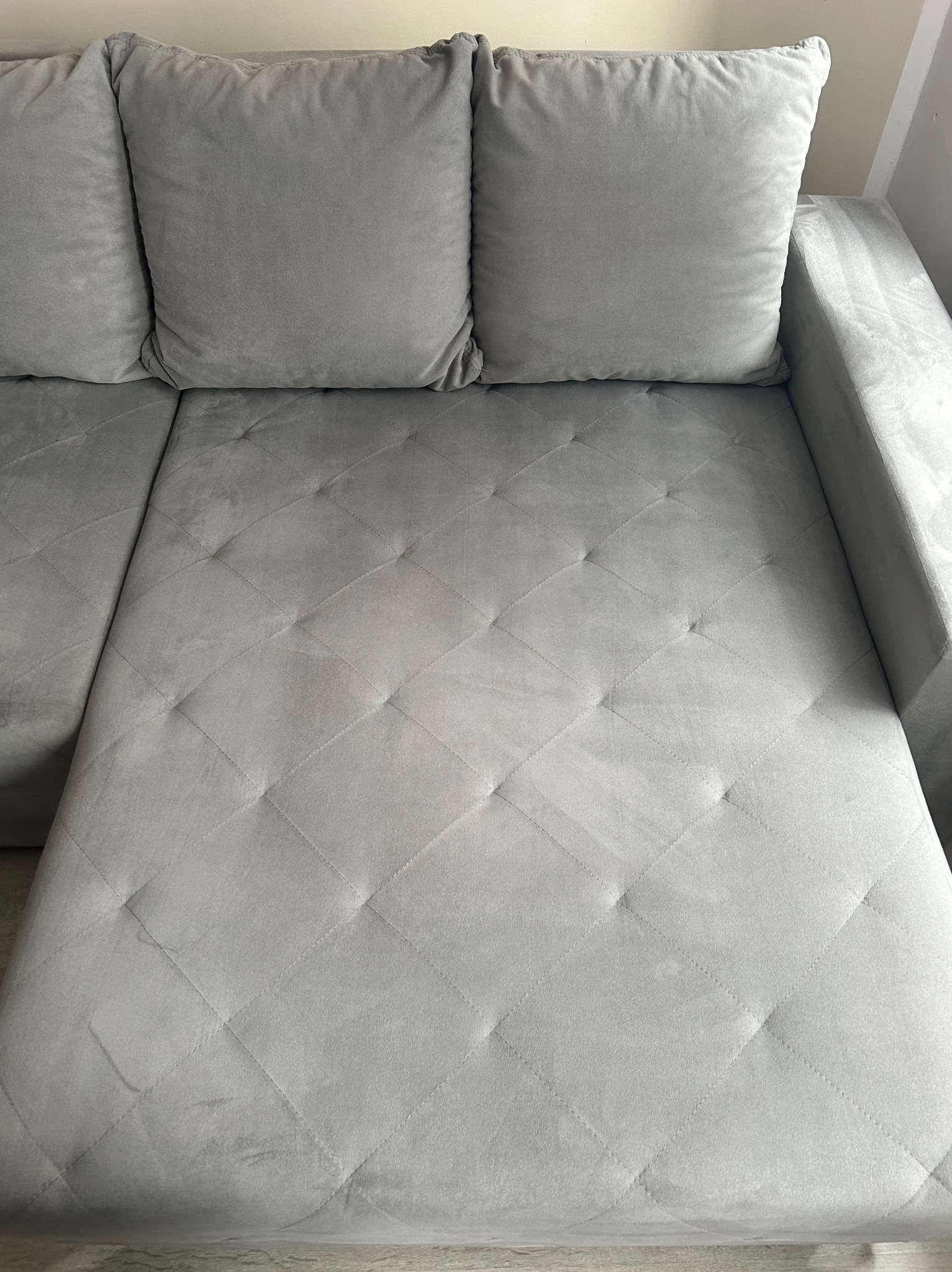 Jasnoszara, welurowa sofa narożna z pikowanym siedziskiem w romby i trzema poduszkami o gładkiej fakturze. Widok z góry, neutralne oświetlenie.