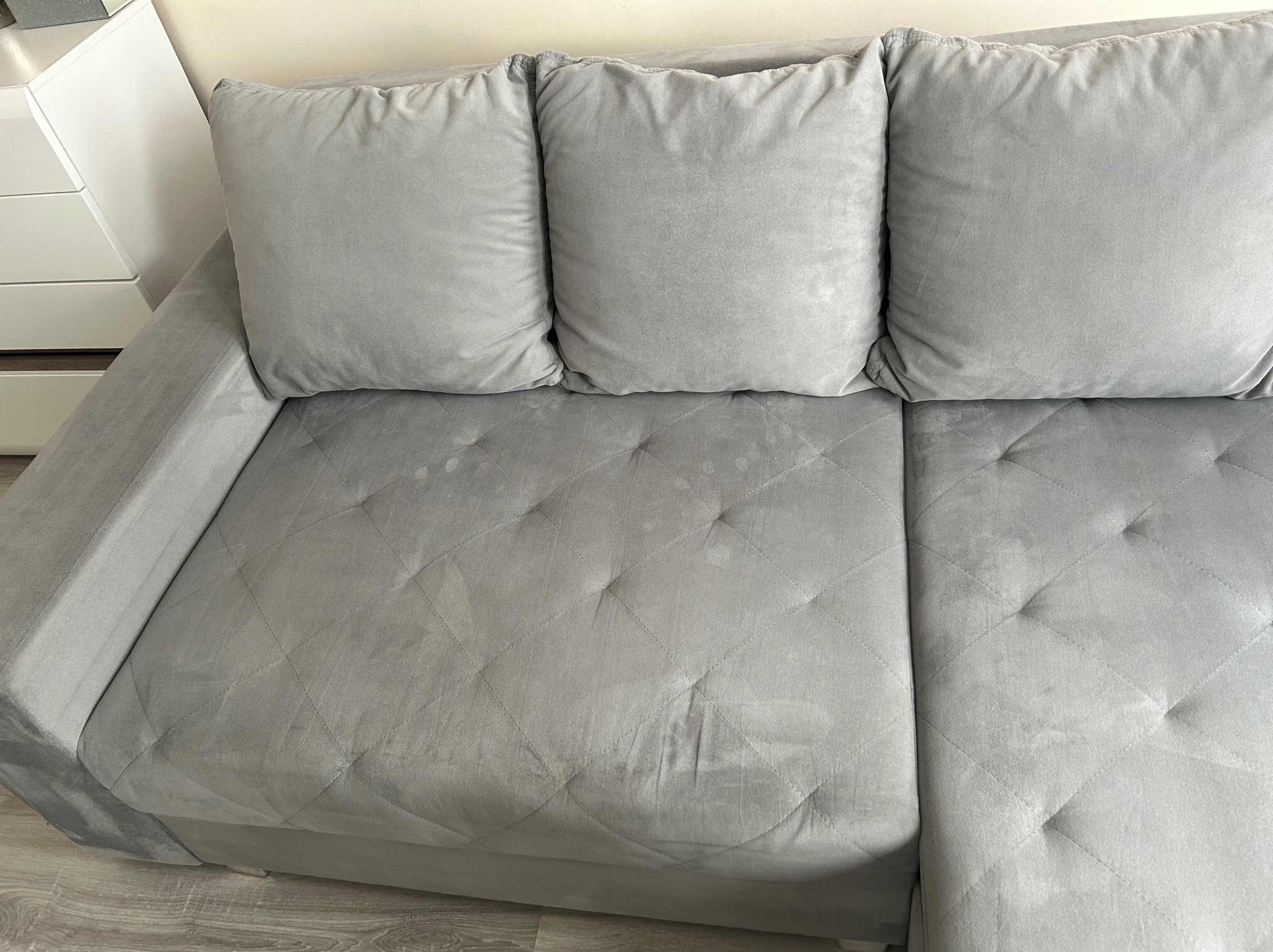 Szara, narożna kanapa z widocznymi plamami na siedzisku i trzema poduszkami oparciowymi. Widok z góry na zabrudzoną tapicerkę.