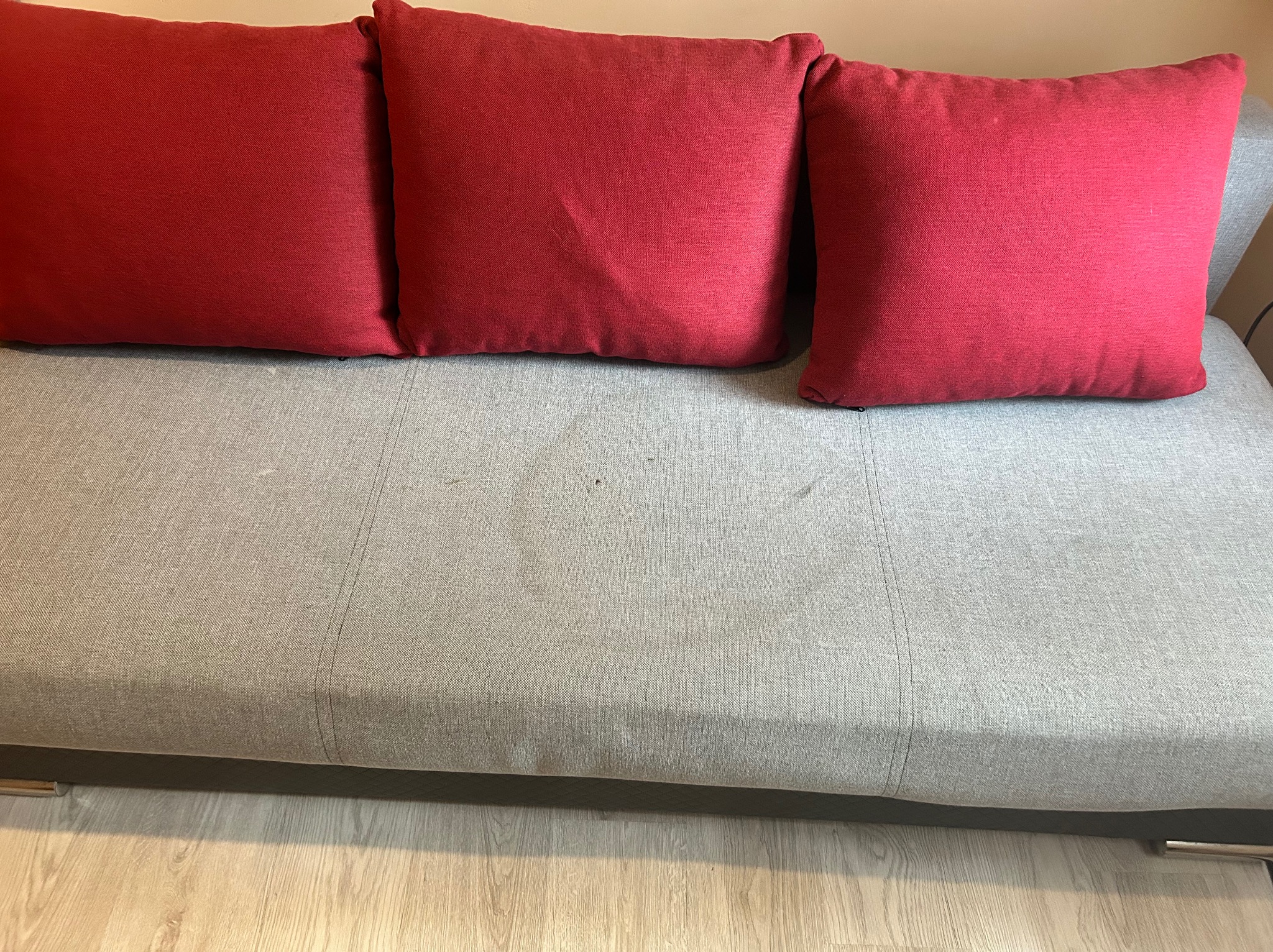 Szara kanapa z trzema czerwonymi poduszkami, widoczne plamy na siedzisku. Zdjęcie 'przed' czyszczeniem tapicerki.