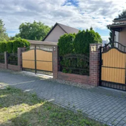 Nowoczesne ogrodzenie z drewnopodobną bramą i furtką, murowane słupki z cegły, dekoracyjne metalowe elementy, wysokie tuje wzdłuż ogrodzenia.