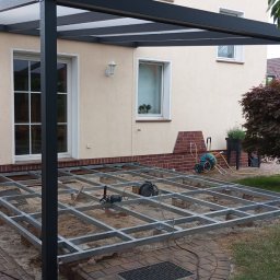 JWFactory - Metalowa konstrukcja tarasu kompozytowego w trakcie montażu, widoczna na tle domu z oknami i zieleni. Etap budowy z narzędziami.