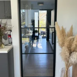 Szklane drzwi z czarną ramą, widok z kuchni na jasny korytarz. Minimalistyczny design, drewniana podłoga, dekoracje z suszonych traw. Nowoczesne wnętrze.