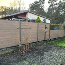 JWFactory - Nowoczesne ogrodzenie kompozytowe w kolorze drewna z metalowymi słupkami, widok z zewnątrz posesji, z elementami ogrodu i domu w tle.