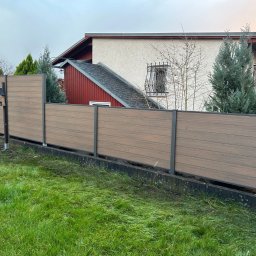 JWFactory - Nowoczesne ogrodzenie kompozytowe w kolorze drewna z metalowymi słupkami, widok z niskiej perspektywy na tle domu i zielonego trawnika.