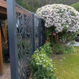 JWFactory - Nowoczesne ogrodzenie metalowe z ażurowym wzorem liści, oddzielające ogród z kwitnącym krzewem. Widoczny fragment altany i trawnik.