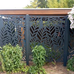 JWFactory - Nowoczesne ogrodzenie panelowe z motywem liści, w kolorze grafitowym, z widocznymi roślinami przed i za nim, oraz fragmentem drewnianej konstrukcji w tle.