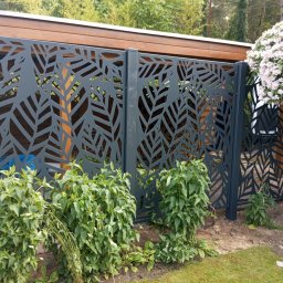 JWFactory - Dekoracyjne, ażurowe ogrodzenie w kolorze antracytu z motywem liści na tle drewnianej konstrukcji i zieleni. Nowoczesny design, podkreślony przez roślinność w ogrodzie.