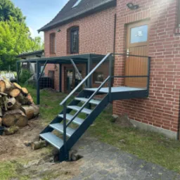 Zewnętrzne, metalowe schody z podestem prowadzą do drzwi w ceglanym budynku. W tle widoczna konstrukcja zadaszenia i ułożone drewno. Słoneczny dzień.