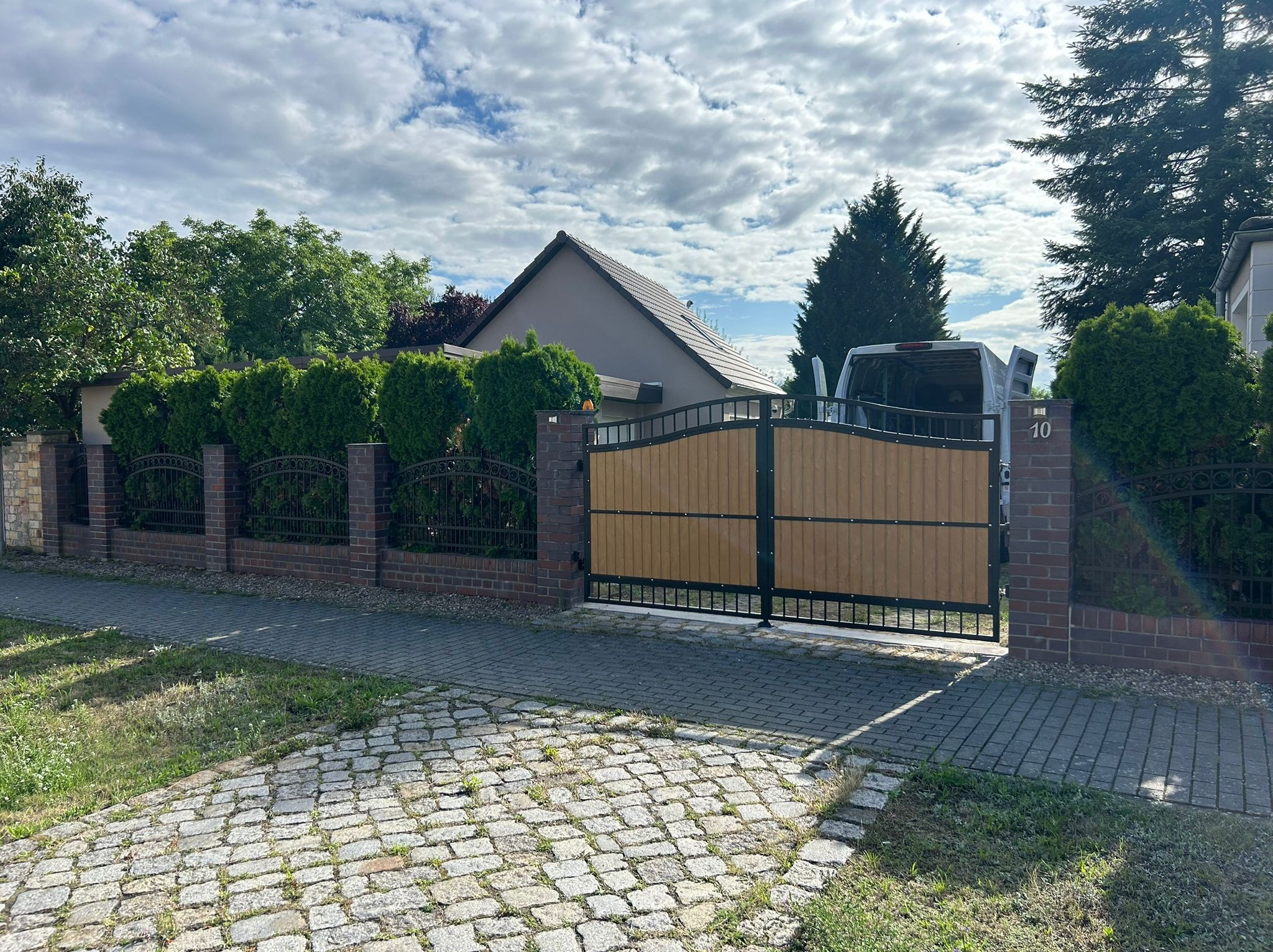 Brama z paneli drewnopodobnych i metalu, wkomponowana w ogrodzenie z cegły i metaloplastyki. W tle dom z ciemnym dachem i zaparkowany samochód dostawczy.